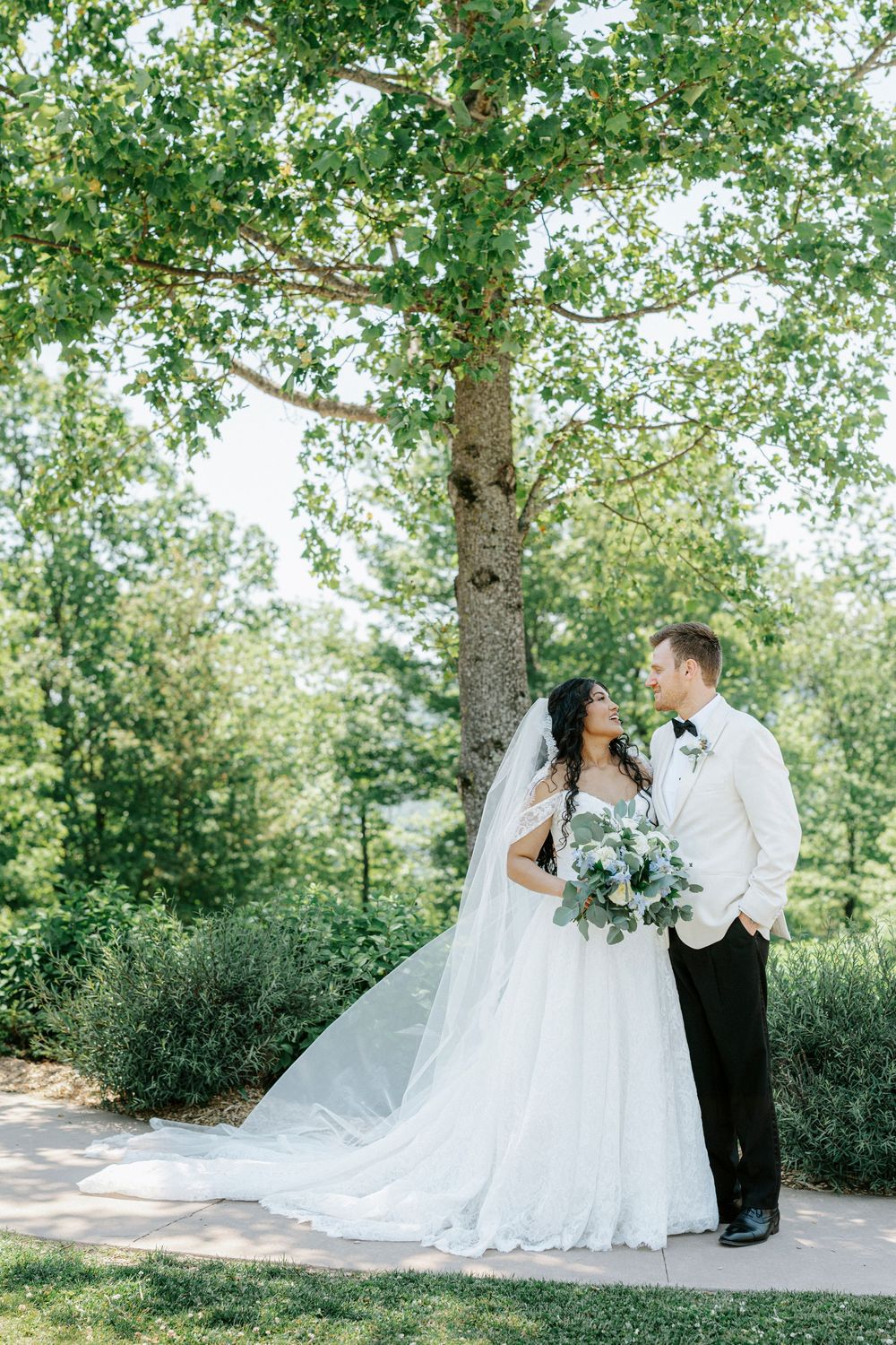 Bride and groom share romantic moment under large tree surrounded by lush greenery at outdoor wedding.