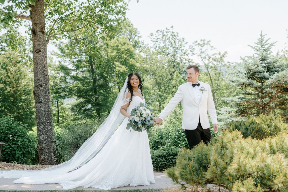 Bride and groom hold hands walking through lush garden, bride wearing cathedral veil and holding bouquet.