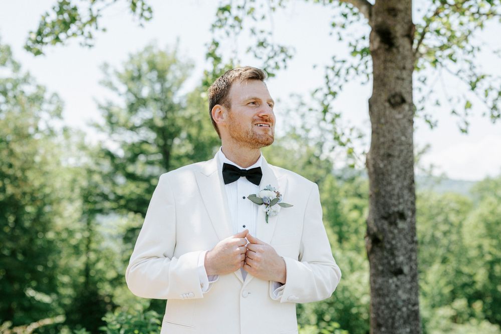 Groom in white tuxedo jacket and black bow tie stands outdoors adjusting jacket near trees.