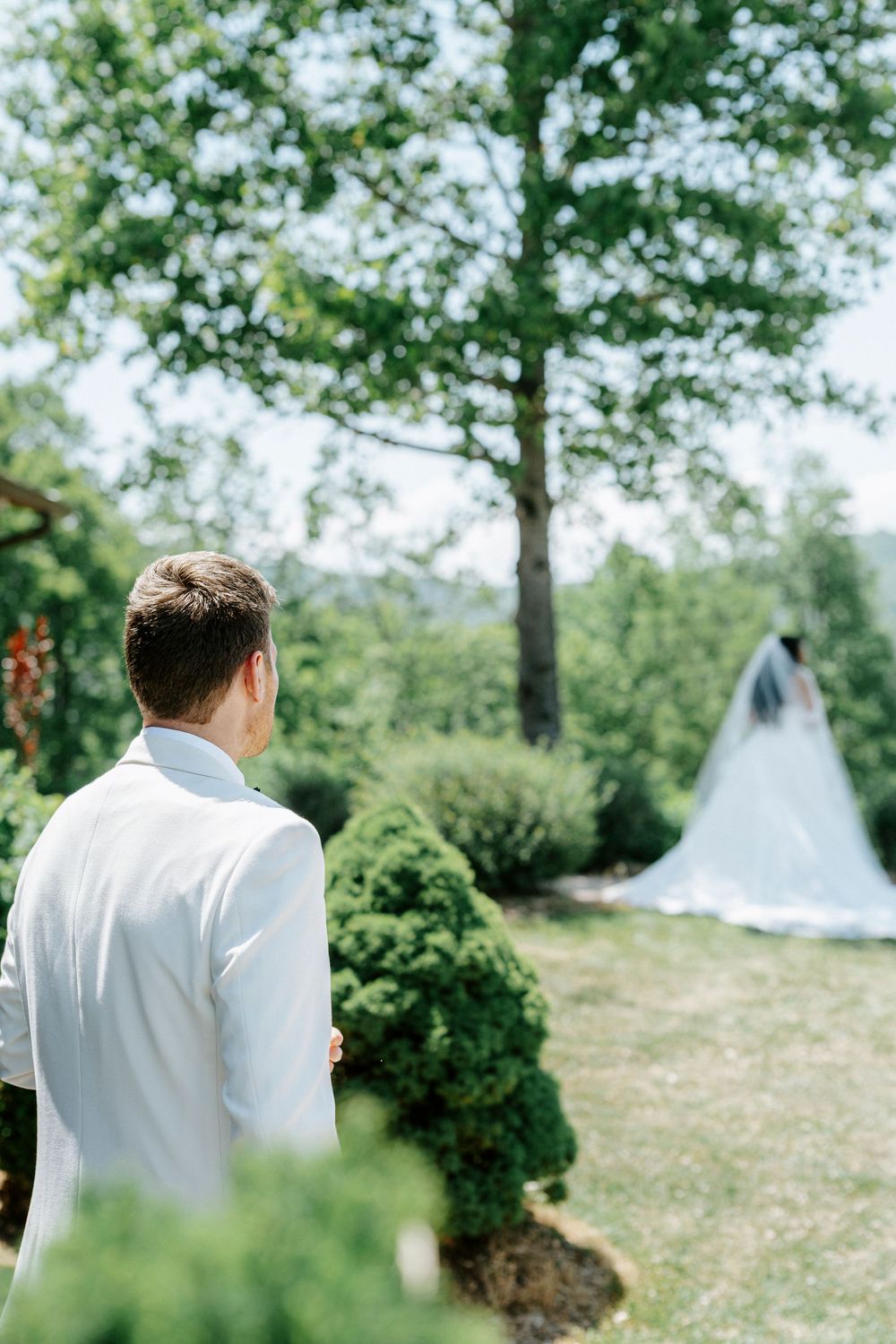 Groom in white suit watches bride in flowing veil across lush green garden on wedding day.
