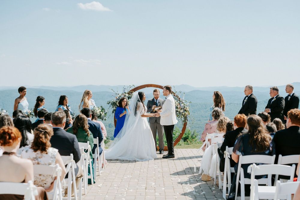 Outdoor wedding ceremony on a mountain terrace with couple at floral arch, guests seated in white chairs under blue sky.