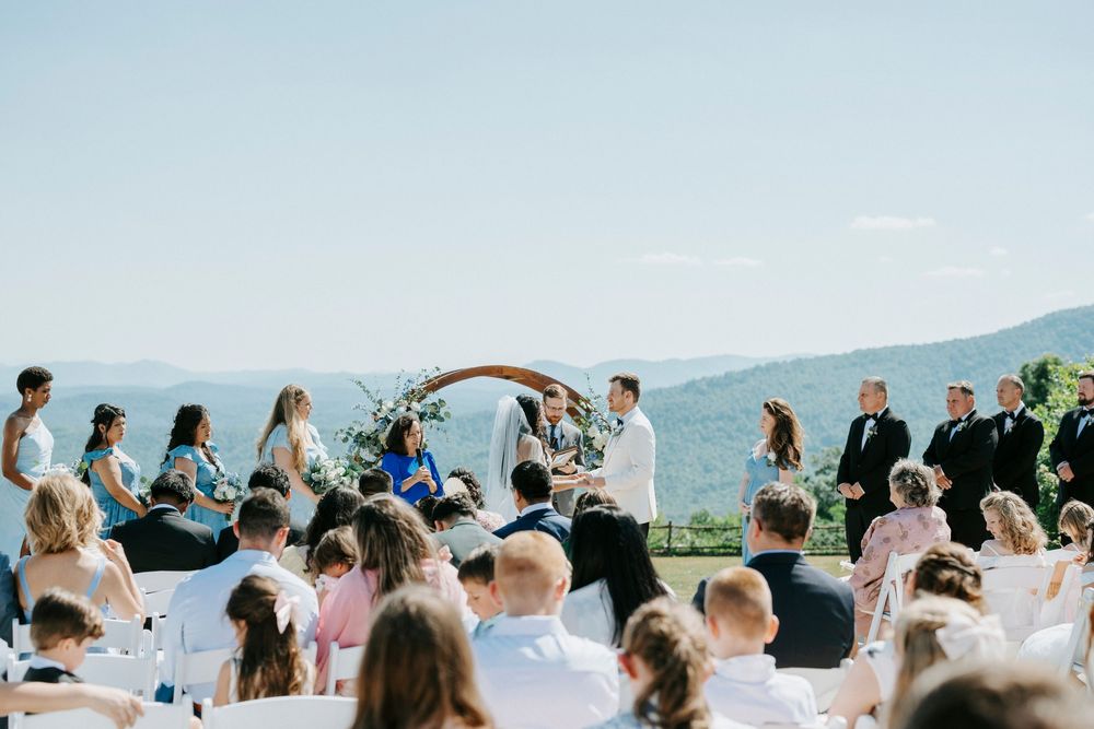 Outdoor wedding ceremony with mountain backdrop, guests seated as couple exchanges vows under floral arch.