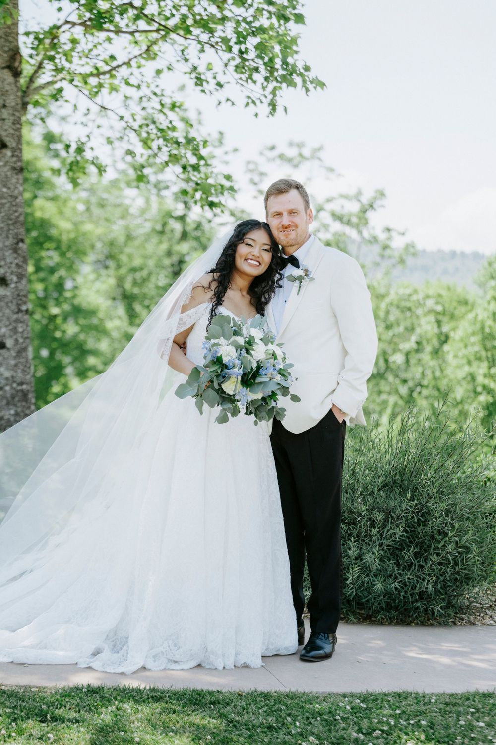 A bride and groom pose outdoors on their wedding day, she in a white ballgown with veil, he in a white tuxedo jacket.
