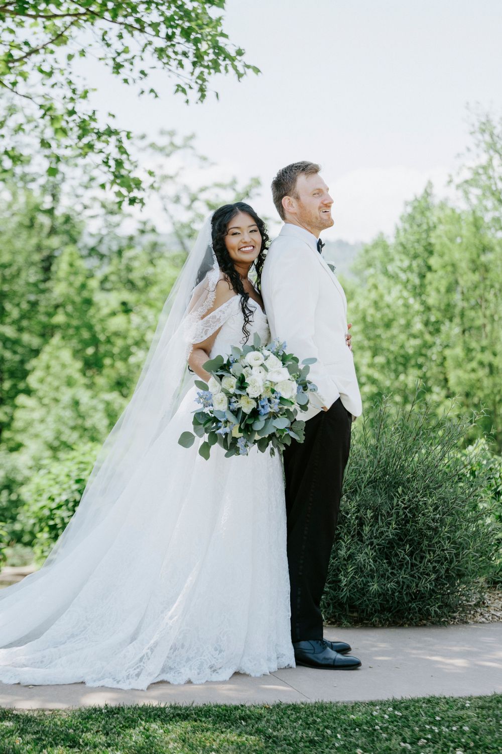 Bride and groom stand back-to-back outdoors, bride holding white and blue bouquet with flowing veil in lush green setting.