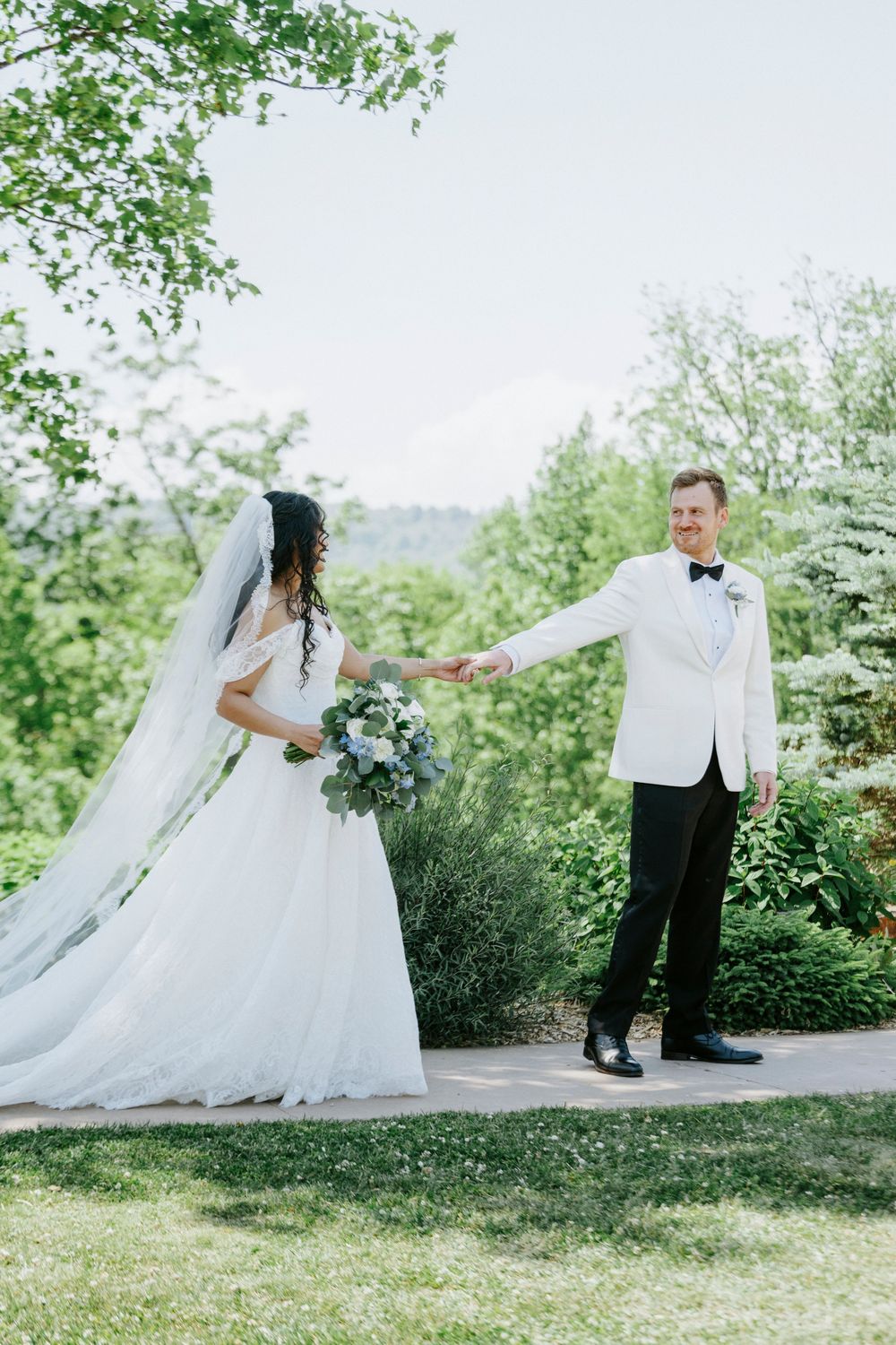 Bride and groom holding hands in a lush garden, groom in white tuxedo jacket, bride in flowing white gown with veil.