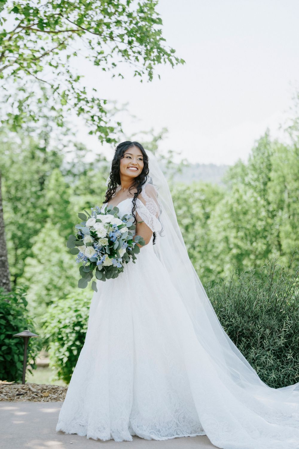 Smiling bride in white lace gown holds bouquet of white flowers and greenery outdoors.