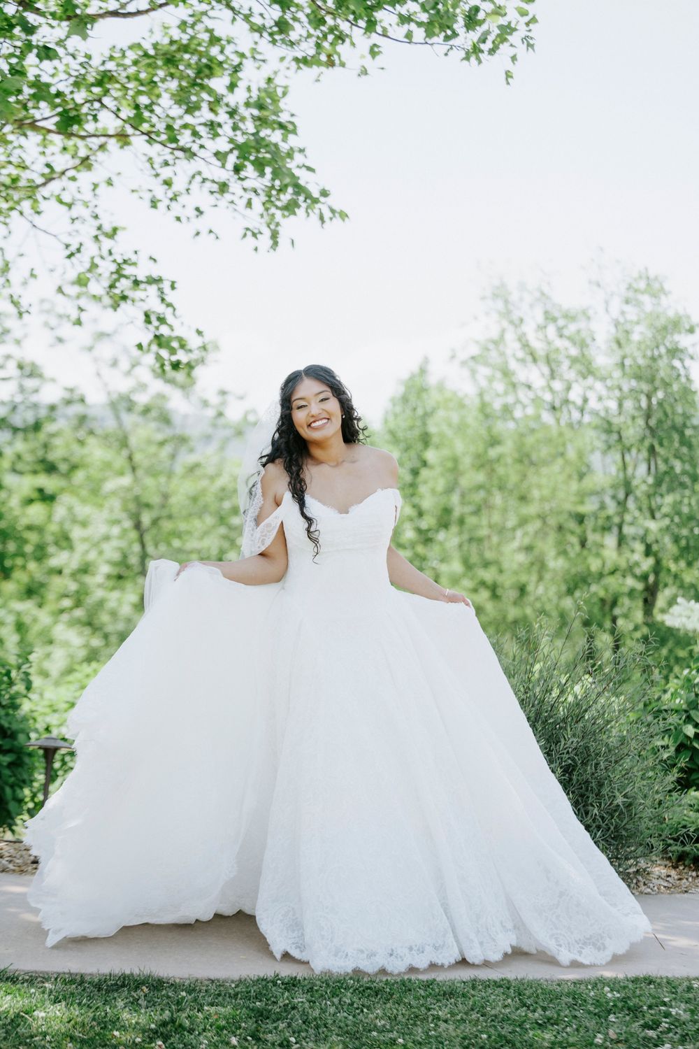 Smiling bride in an off-shoulder white ballgown twirling outdoors surrounded by green trees on her wedding day.