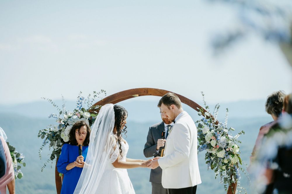 Groom reads vows to bride at outdoor mountain wedding ceremony beneath wooden circular floral arch.