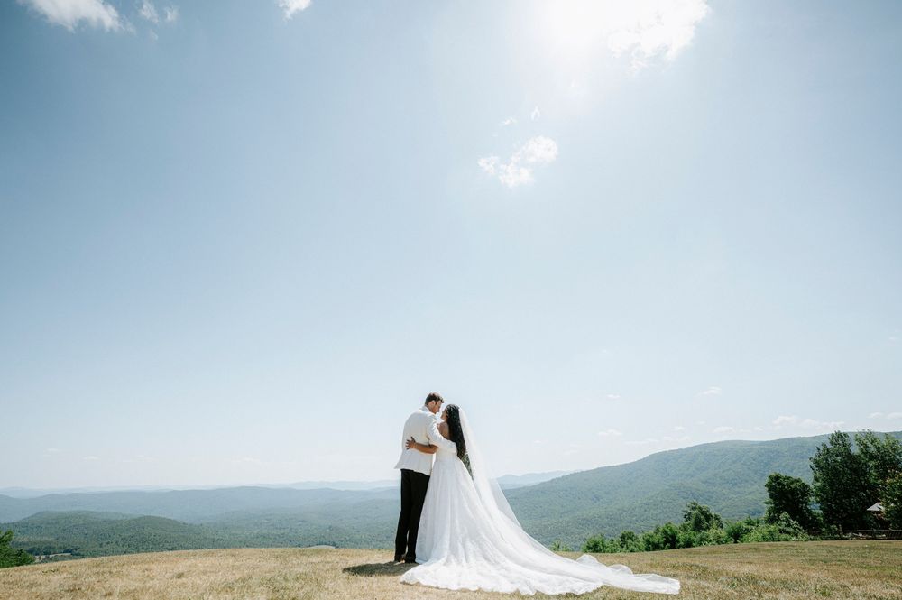 Bride and groom embracing on a hilltop with stunning mountain views and flowing wedding dress train on a sunny day.