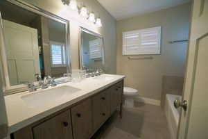 Modern bathroom with double sink vanity, large mirrors, white countertop, toilet, and bathtub with neutral tile flooring.