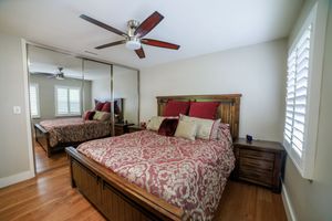 Cozy bedroom with queen bed, red bedding, wood furniture, ceiling fan, mirrored closet doors, and plantation shutters.