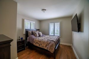 Cozy bedroom with hardwood floors, dark wood furniture, wall-mounted TV, and two windows with white shutters.