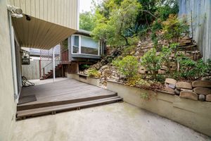 Wooden deck with steps leads to a hillside home surrounded by lush greenery and a terraced stone retaining wall.