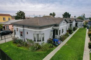 Aerial view of a gray single-story home with brown roof, green lawn, and mature trees in a residential neighborhood.