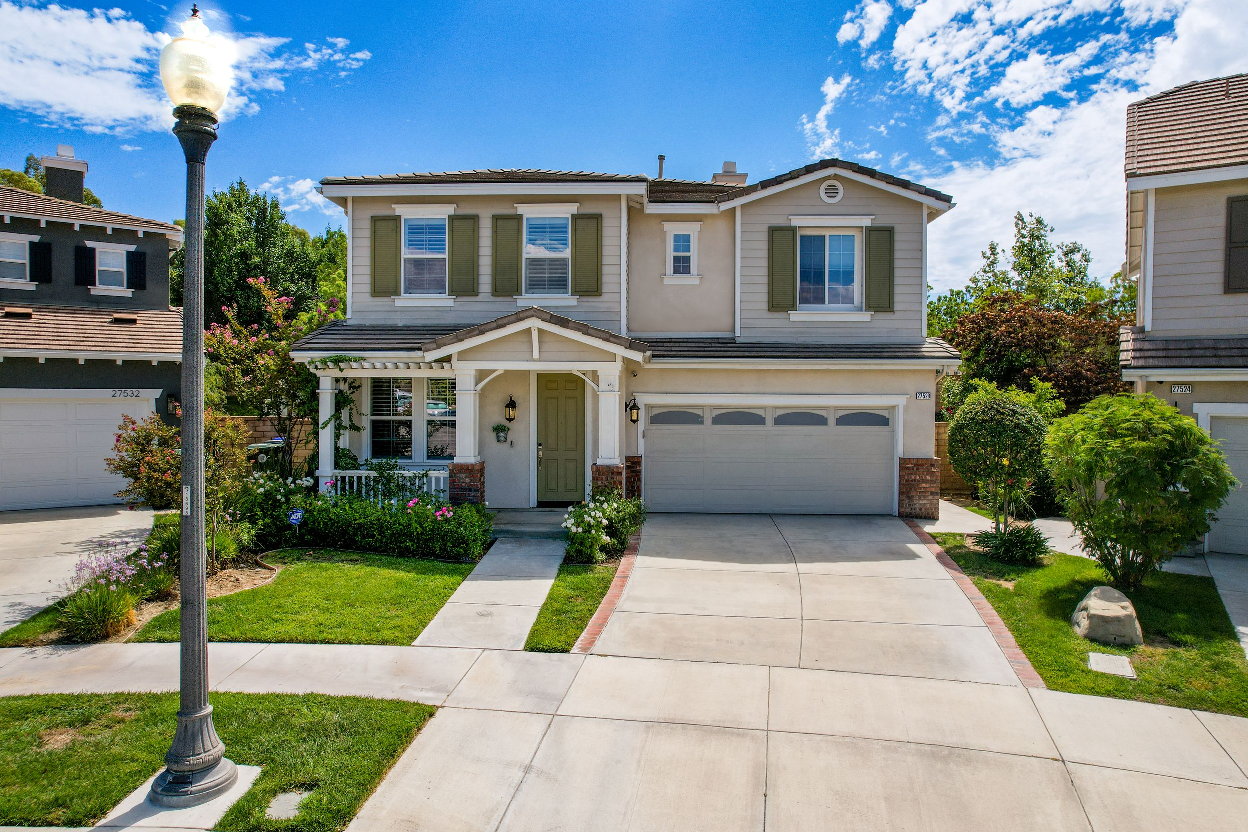 Two-story suburban home with beige exterior, green shutters, attached garage, manicured lawn, and a street lamp in a sunny neighborhood.
