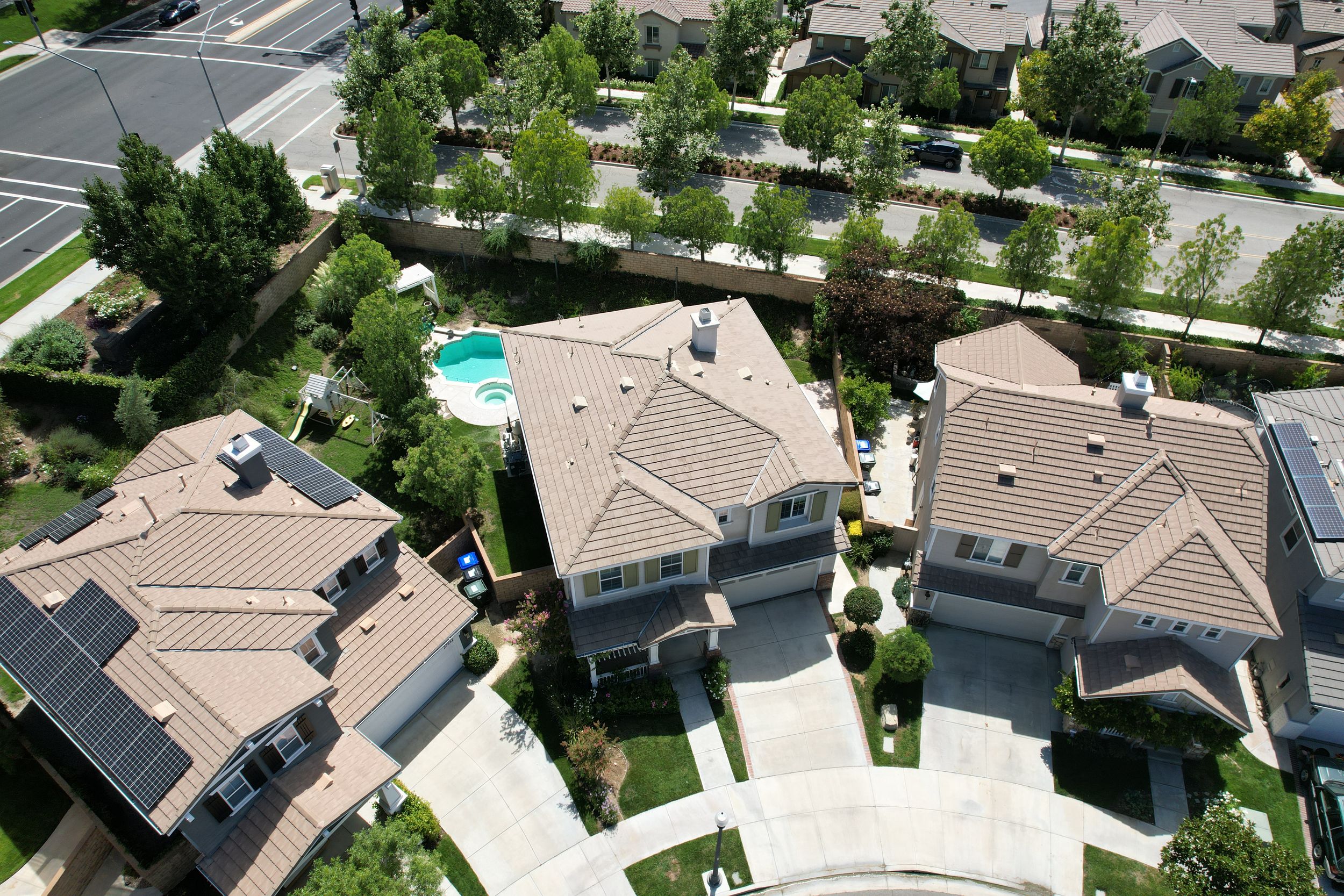 Aerial view of suburban homes with tile roofs, solar panels, manicured lawns, and a backyard pool in a residential neighborhood.