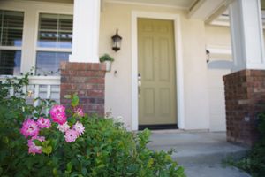 Front porch of a home with olive green door, brick accents, and pink roses blooming in lush green bushes.