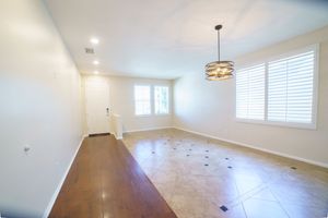 Bright empty room with hardwood and tile floors, white walls, plantation shutters, and an elegant hanging light fixture.