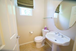 Small bathroom with pedestal sink, round mirror, toilet, window with roman shade, and neutral tile flooring.
