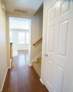 Bright hallway with hardwood floors, white six-panel door, staircase with wood railing, leading to sunlit room.