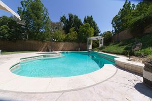 Backyard swimming pool with spa, pergola, and lush green trees on a sunny day.