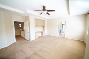 Empty living room with beige carpet, ceiling fan, built-in desk, and open floor plan in a new home.