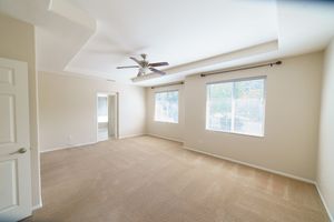 Bright empty living room with beige carpet, ceiling fan, tray ceiling, and large windows allowing natural light.