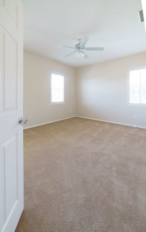 Empty bedroom with beige carpet, white ceiling fan, two windows, and neutral walls in a move-in ready home.