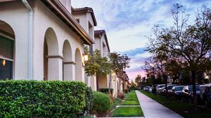 Suburban townhomes with arched facades, lush hedges, and a tree-lined sidewalk at dusk in a quiet neighborhood.