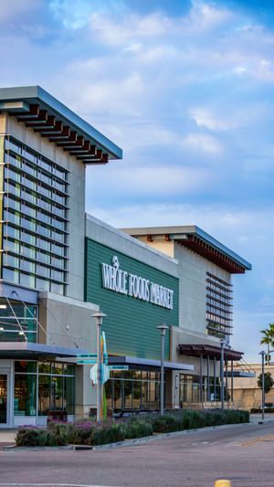 Whole Foods Market storefront exterior with modern architecture, green facade, and cloudy blue sky in background.