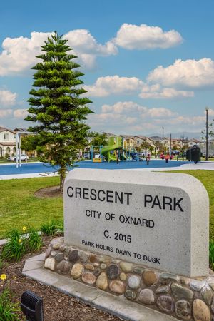 Stone sign for Crescent Park, City of Oxnard, established 2015, with playground and blue sky in background.