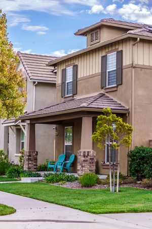 Two-story suburban home with tan stucco exterior, brown shutters, covered porch with teal Adirondack chairs, and well-manicured lawn.