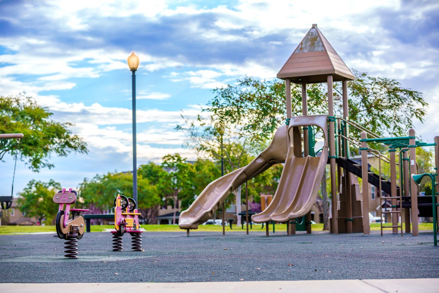 Children playing on a large playground structure with slides in a spacious park under a partly cloudy sky.