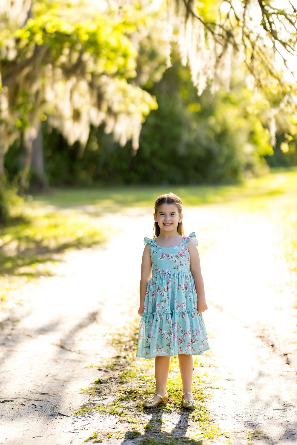 Young girl in floral turquoise dress standing on sunlit path under Spanish moss-draped trees.