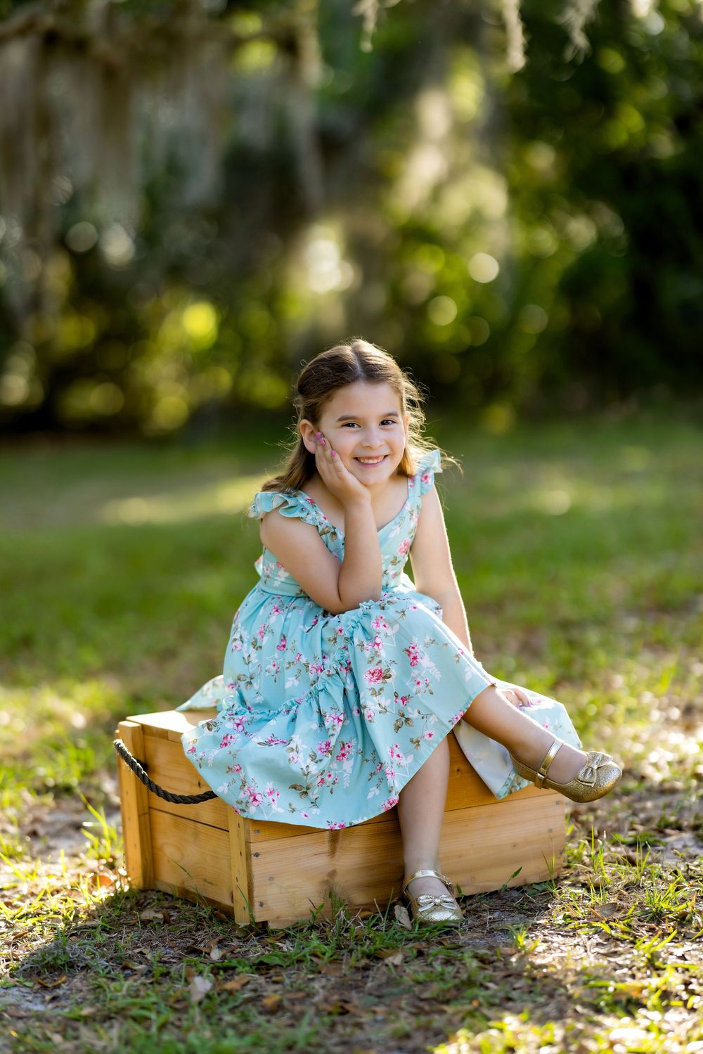 Young girl in floral teal dress sitting on wooden crate outdoors, smiling with hand on cheek in lush green park setting.