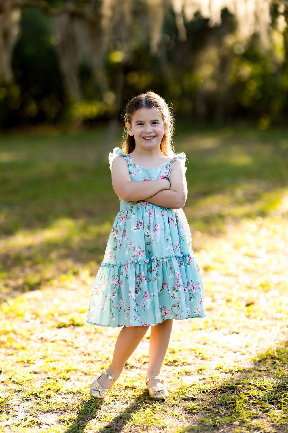 Young girl in teal floral dress stands with arms crossed, smiling outdoors in sunlit park.