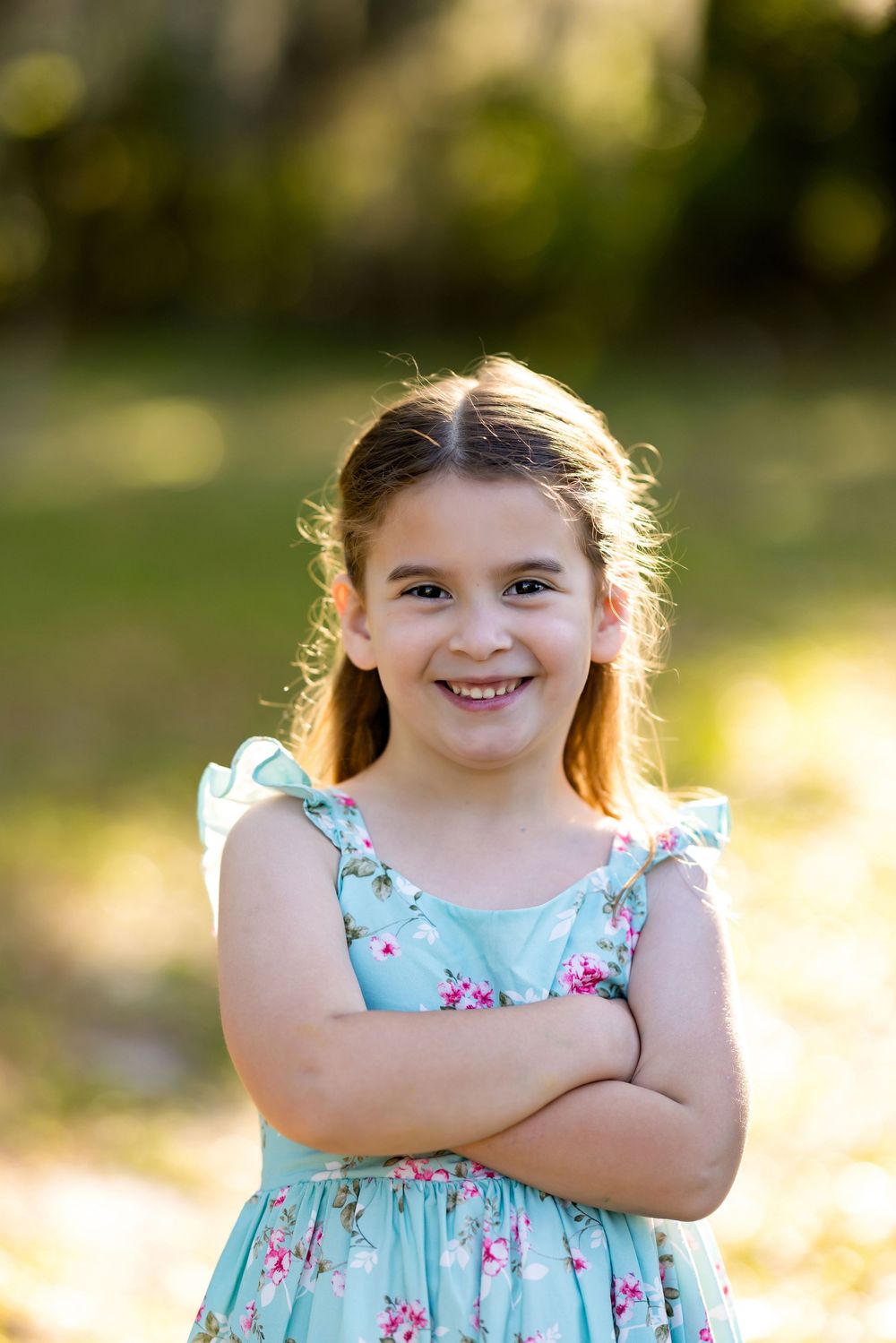 Smiling young girl in a floral teal dress standing outdoors with arms crossed in a sunny park setting.