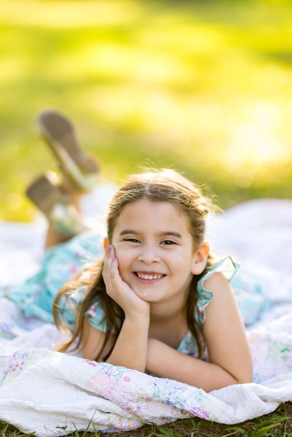Smiling young girl in a blue floral dress lying on a blanket in a sunny park, resting her chin on her hand.