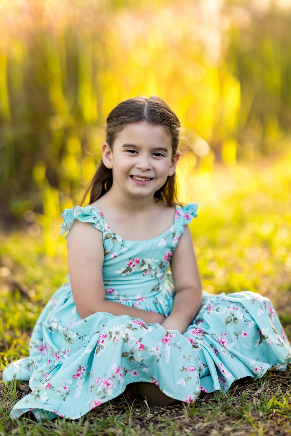 Young girl in teal floral dress sitting on grass, smiling outdoors in golden sunlight.