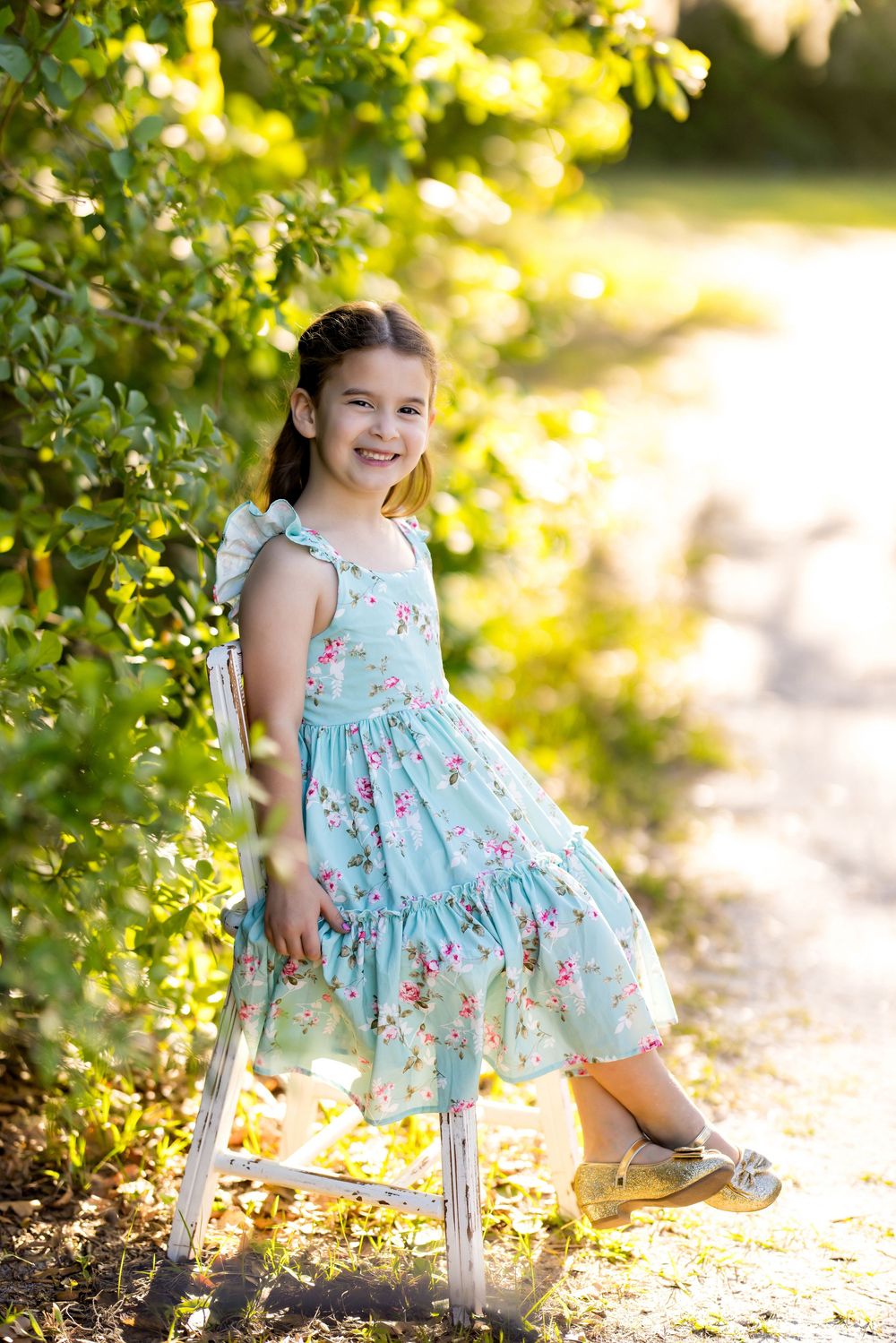 A smiling young girl in a floral mint dress poses by green bushes outdoors in golden sunlight.