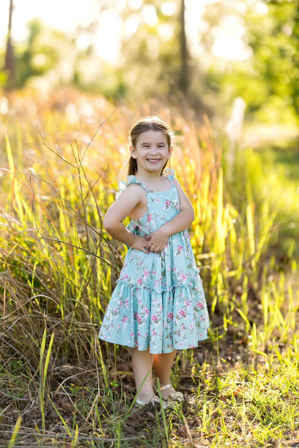 Happy young girl in teal floral dress poses among tall grass in golden sunlight outdoors.
