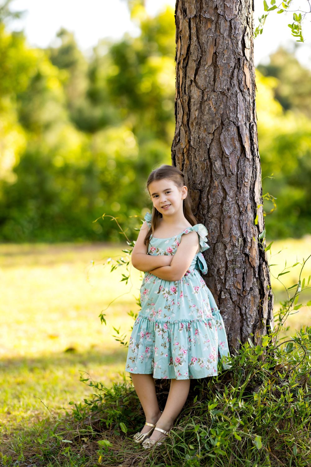 Young girl in floral dress leaning against tree trunk in sunny park, arms crossed, smiling.