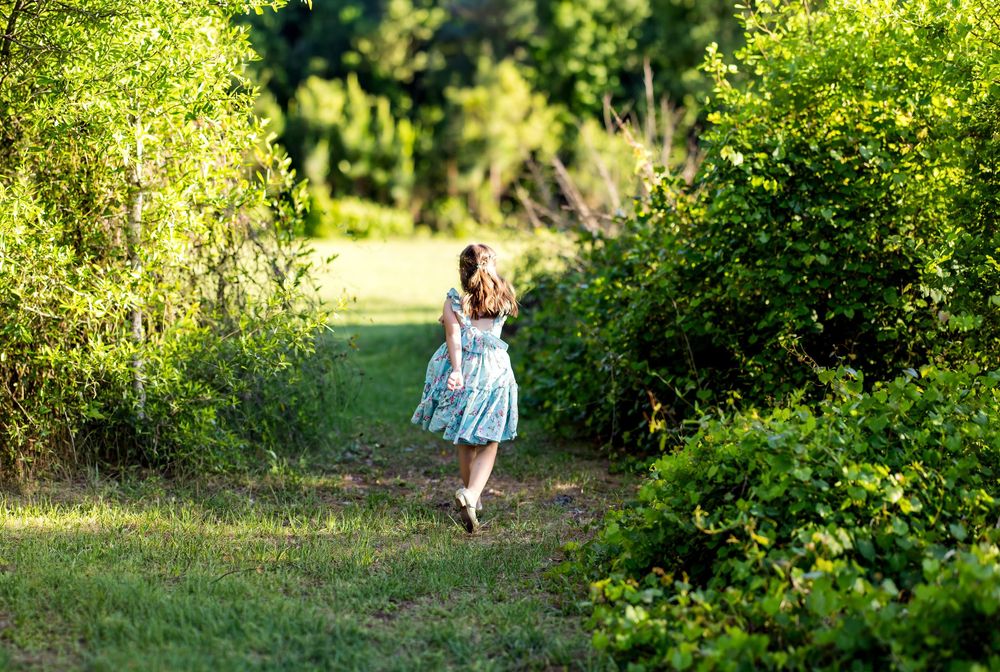A young girl in a floral dress runs along a lush green forest path surrounded by trees.