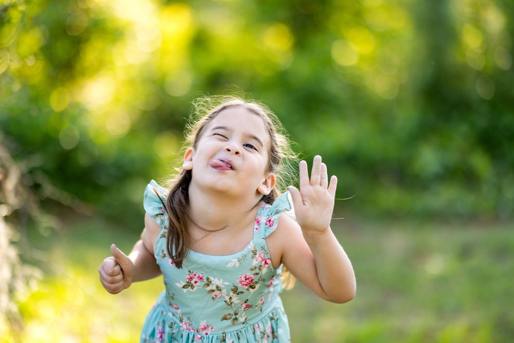 A cheerful little girl in a floral dress waves and smiles outdoors in a sunny green garden.