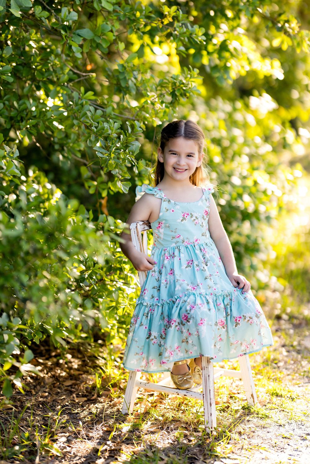 Young girl in floral teal dress smiling outdoors surrounded by lush green trees in warm sunlight.