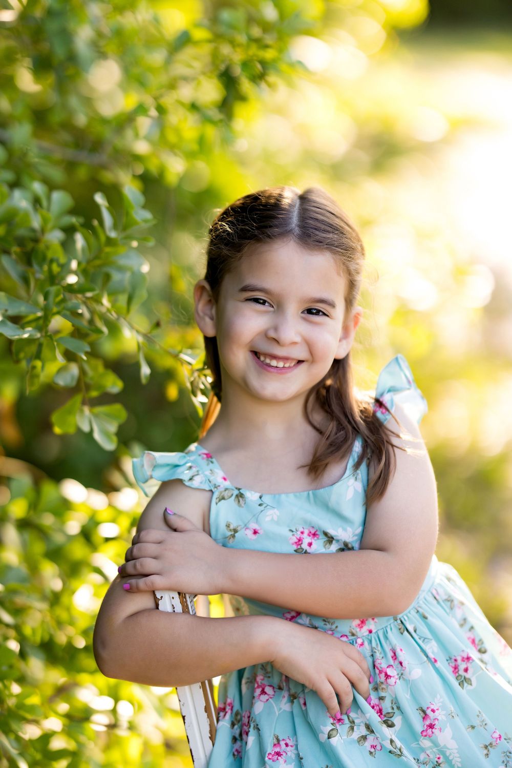 Smiling young girl in a floral teal dress posing outdoors in sunlit garden with lush green foliage background.