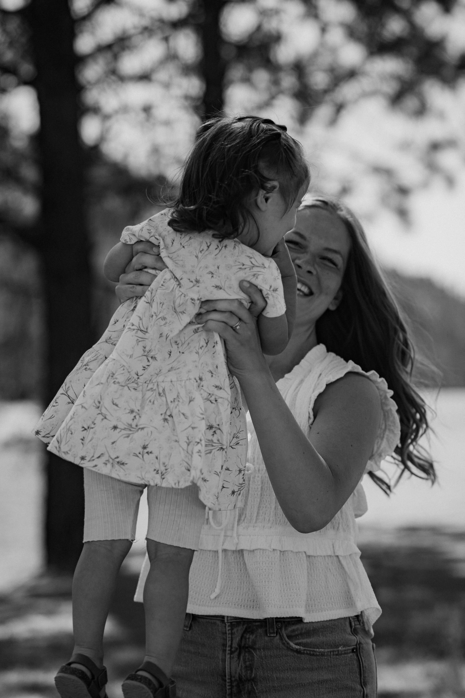 Black and white photo of a smiling woman holding a young girl in a floral dress outdoors.