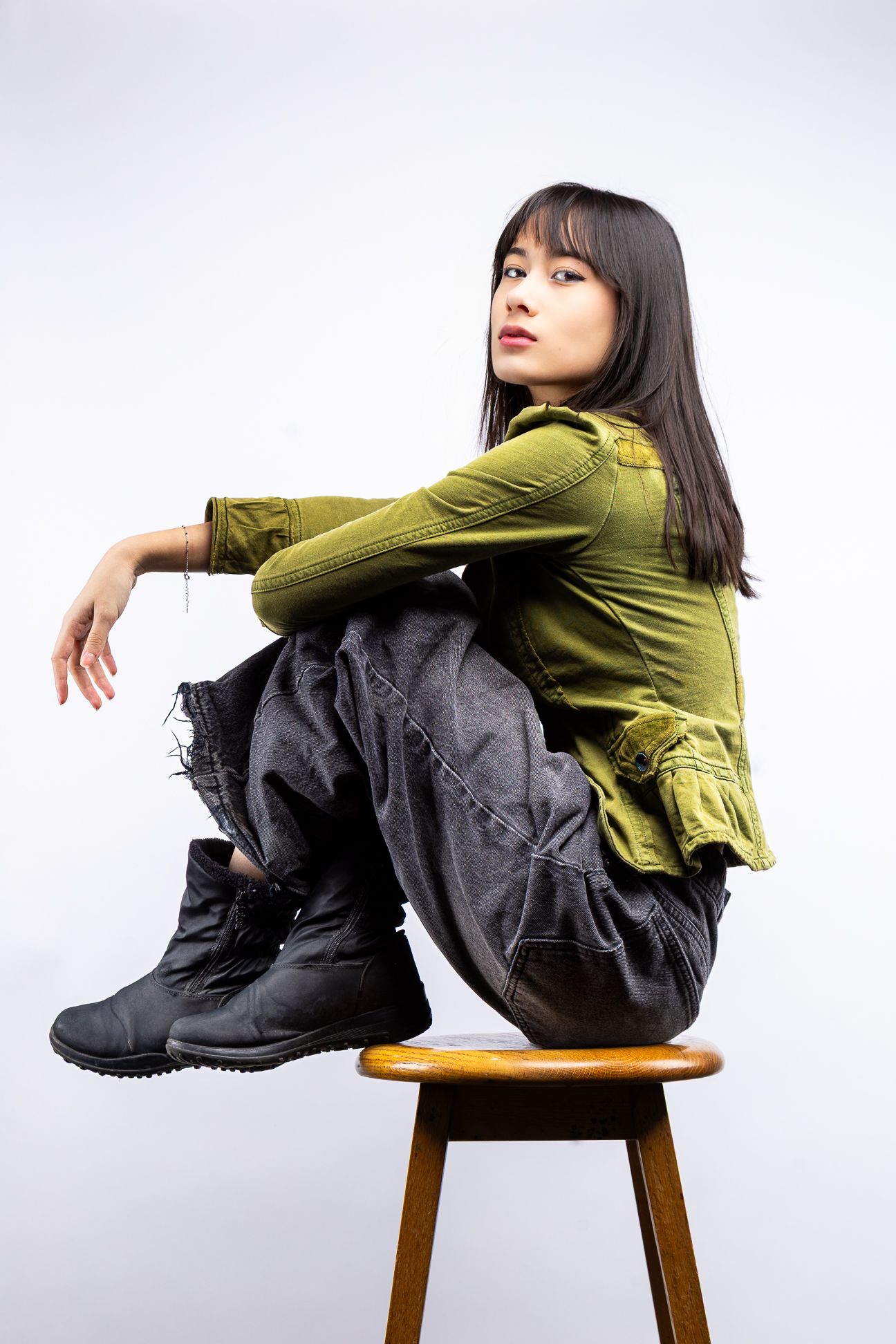 Girl in front of a white backdrop in a green button up blouse, black jeans and shoes, sitting on a stool looking directly at camera