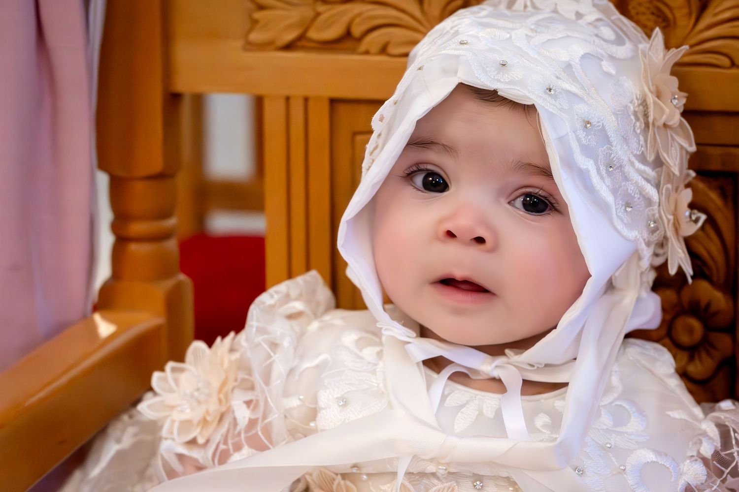 Adorable baby in white lace christening gown and bonnet sitting in wooden chair during baptism ceremony.