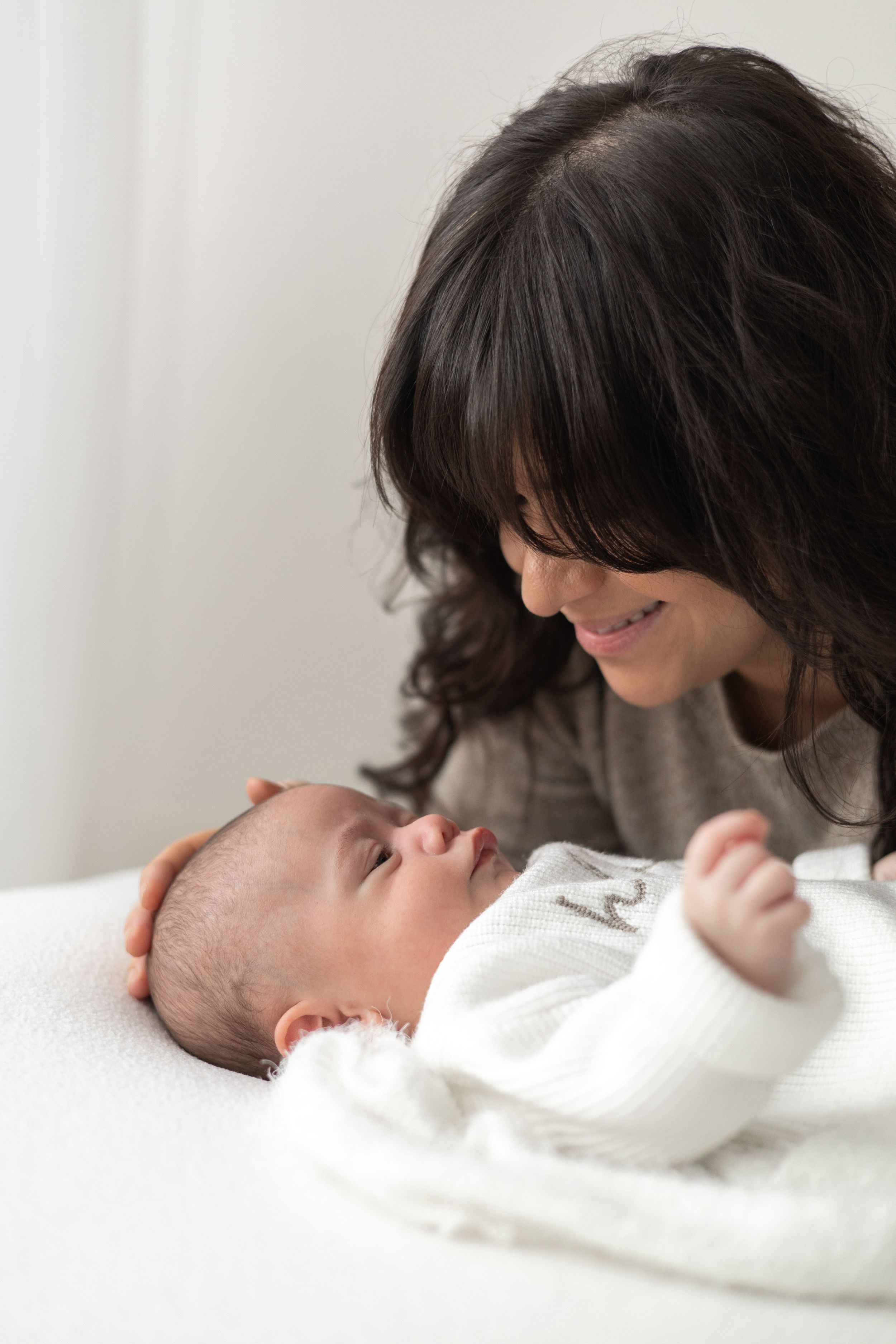 Mother lovingly gazing at her newborn baby wrapped in white blanket lying on bed older baby photographer Nottingham newborn session